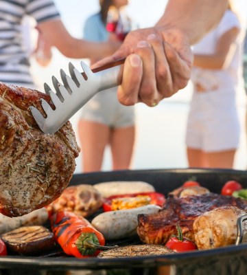 A person holds steak with metal tongs over a barbecue